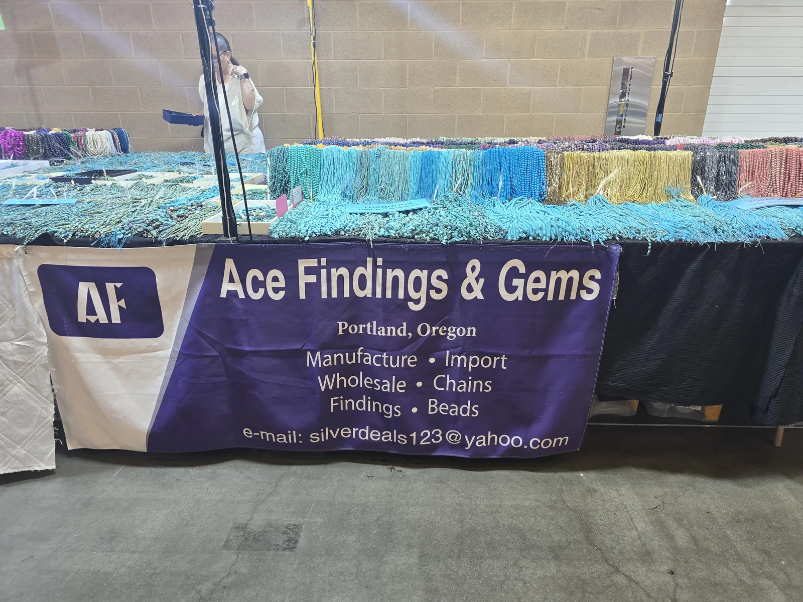 Ace Findings and Gems 展位,深紫色横幅写着 Portland Oregon、Manufacture Import Wholesale、Chains Findings Beads,桌面挂满整排绿松石与彩色碎石珠串