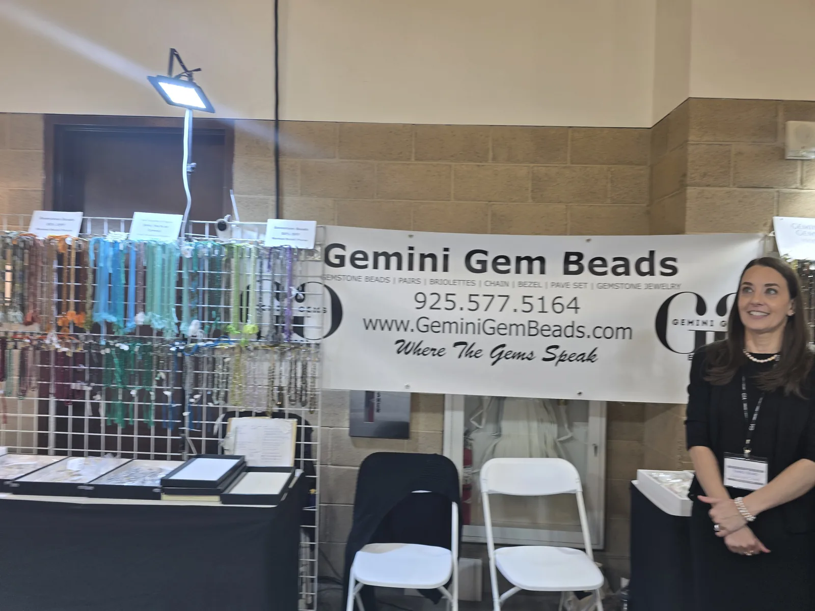 Gemini Gem Beads 展位,横幅写有品牌名与 GEMSTONE BEADS、PAIRS、BRIOLETTES、CHAIN、BEZEL、PAVE SET 等品类,左侧网格架挂满彩色宝石珠串,展商站在右侧