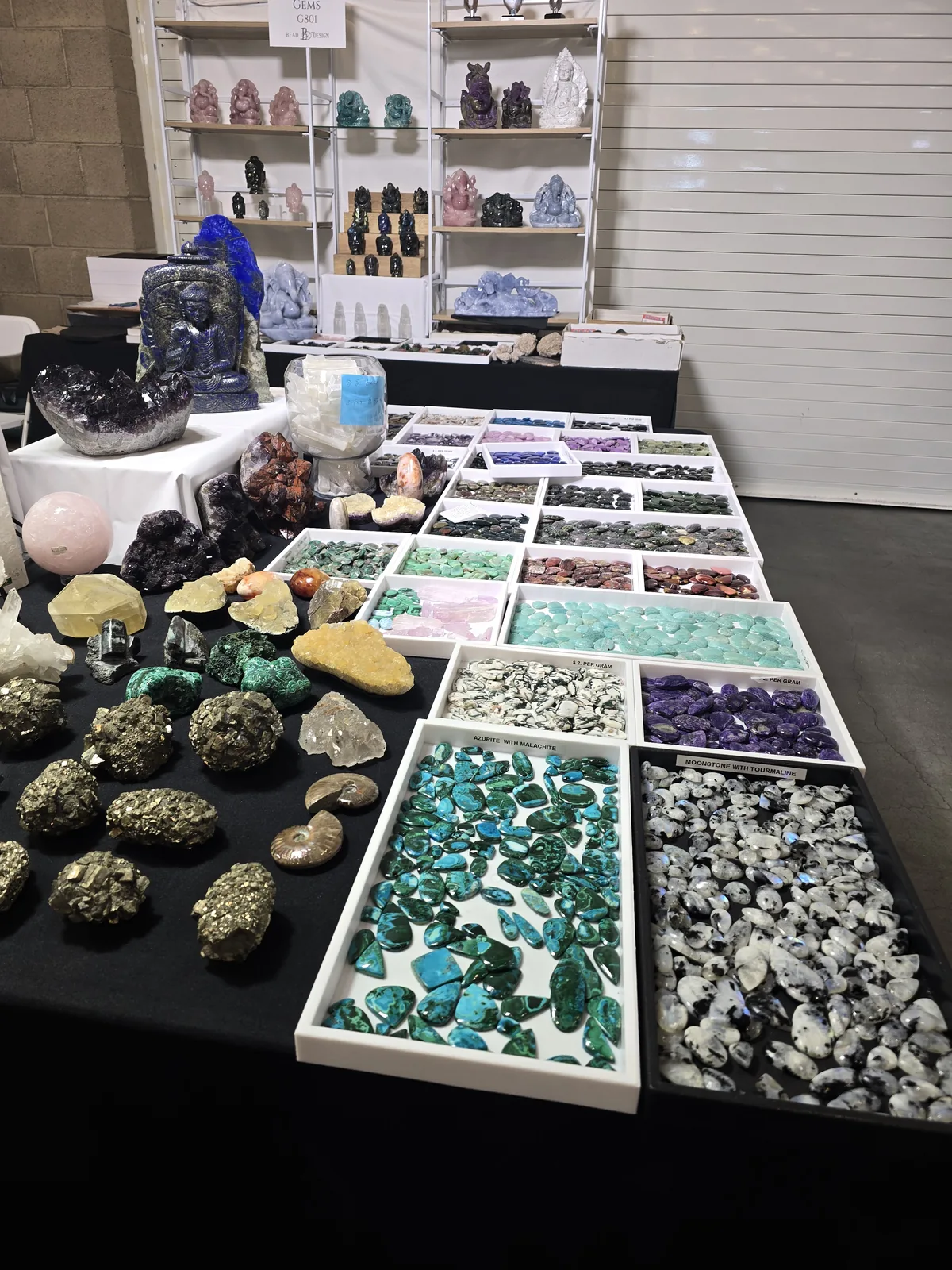 展位桌上摆满黄铁矿原石、孔雀石与水晶,后方白色分格盘装着各色打磨扁石,盘边贴着 PER GRAM 标价签,货架上陈列石雕摆件