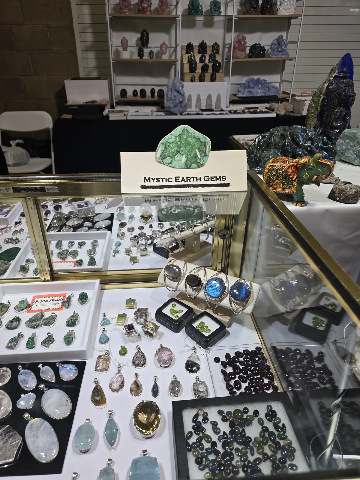 Mystic Earth Gems 展位的金色玻璃柜,柜内陈列银镶宝石戒指与吊坠,柜面上摆着月光石、拉长石手镯与散装石榴石、欧泊