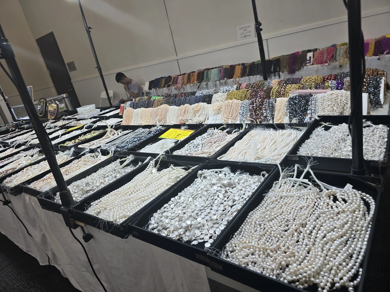 Top Pearl 展位整桌淡水珍珠,黑色浅盘里按形状分装白色圆珠、异形巴洛克珠、扁片珠,后墙挂着一排排彩色珠串