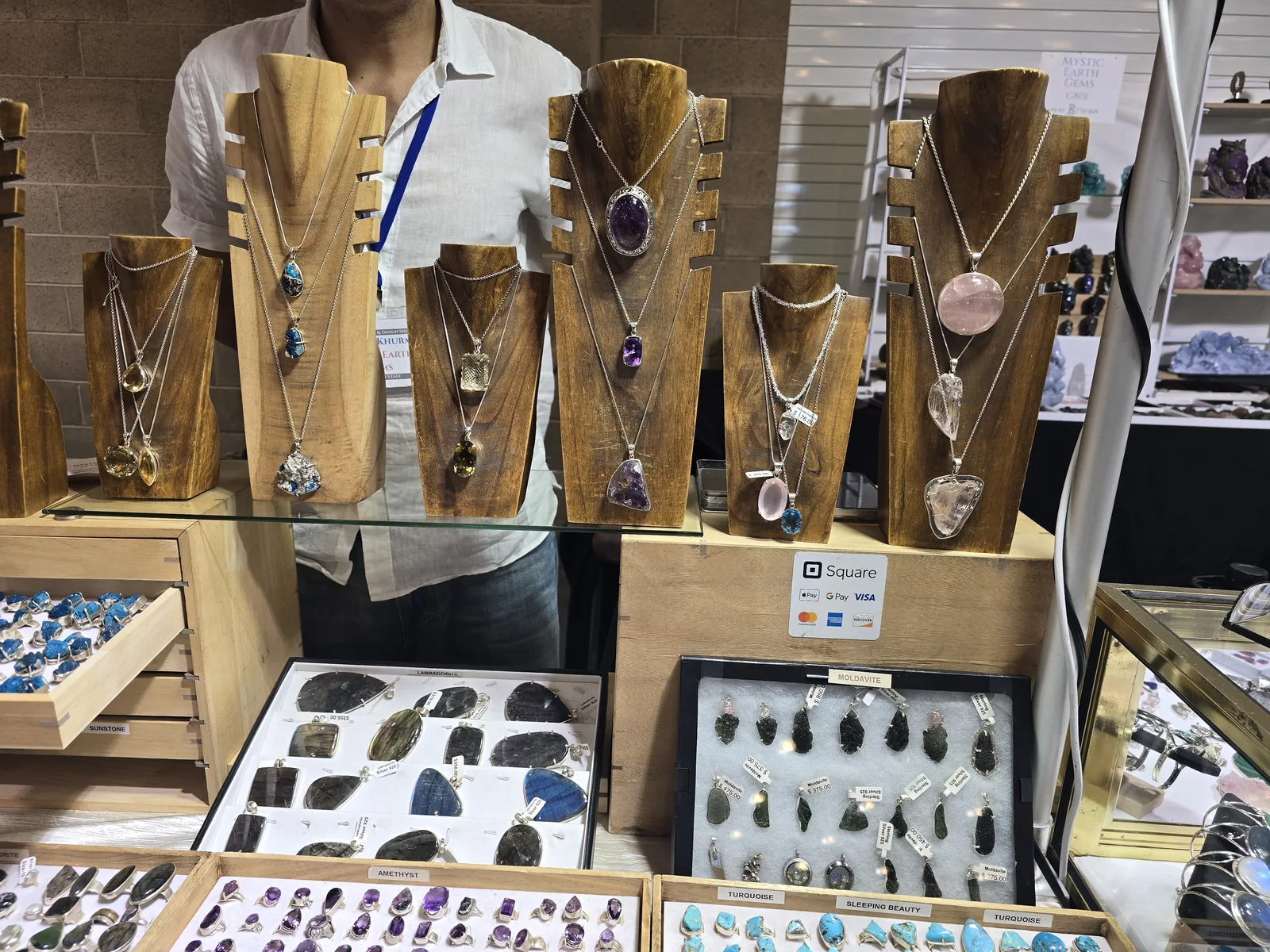 展位木质颈托陈列多条银链宝石吊坠项链,紫水晶、粉水晶、拉长石款式,下方抽屉盘装绿松石戒指与摩尔达石吊坠,柜上贴着 Square 支付标识