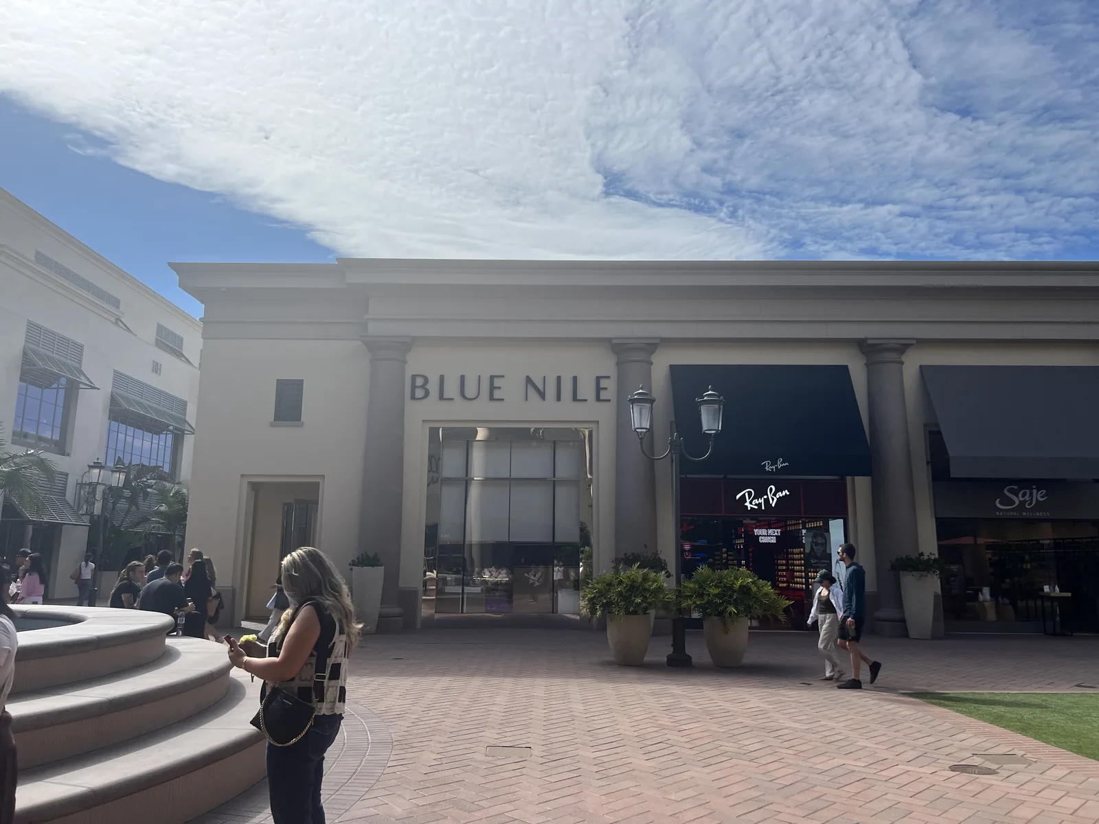 Blue Nile 门店在 Fashion Island 露天广场的外景,米色石材立面、落地玻璃,门前喷泉与人流,旁边是 Ray-Ban 与 Saje
