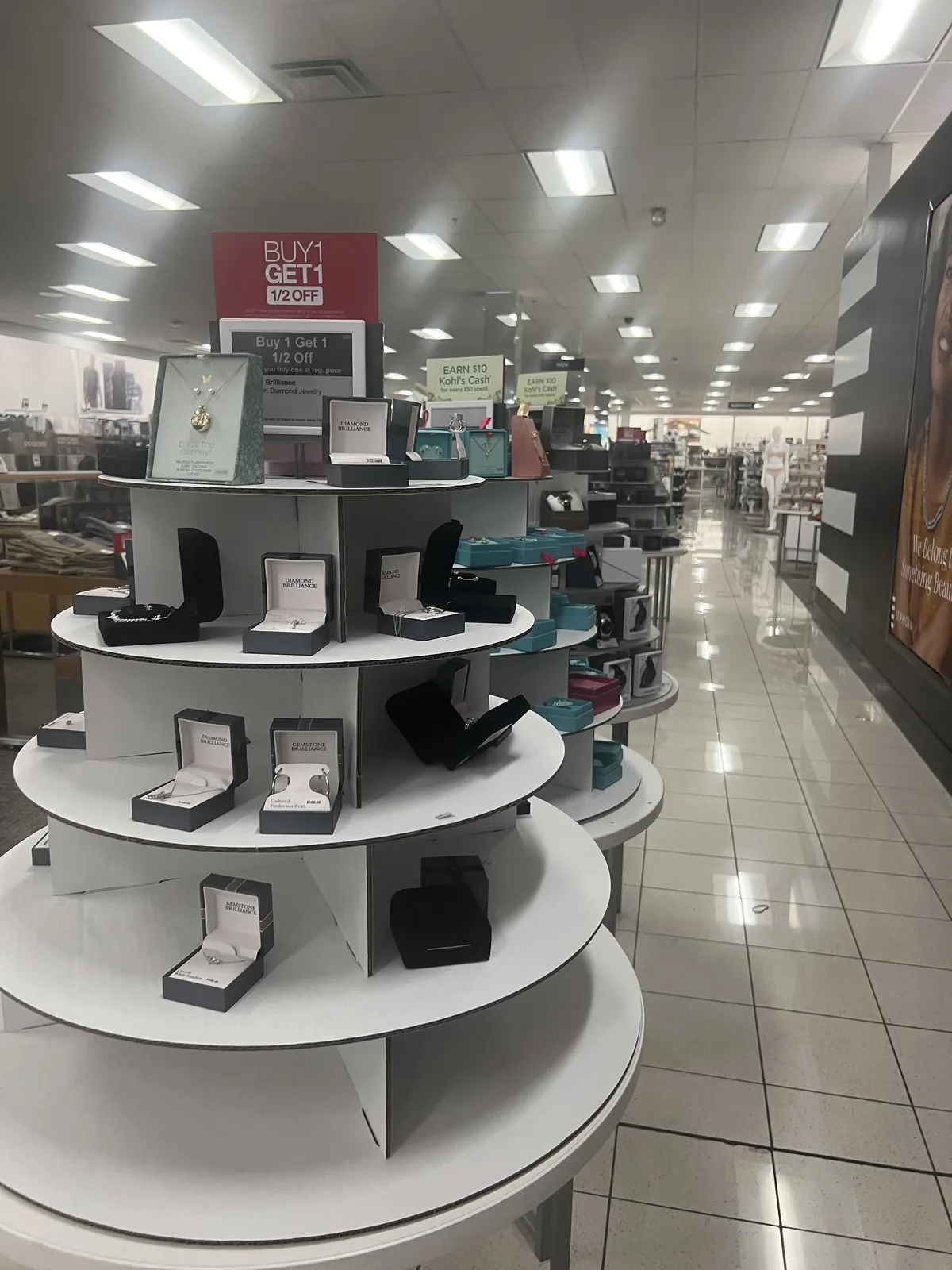 Kohl's 精品珠宝陈列塔,红色 BUY 1 GET 1 1/2 OFF 牌,Diamond Brilliance 盒装吊坠耳钉与 EARN 10 Kohl's Cash 促销牌